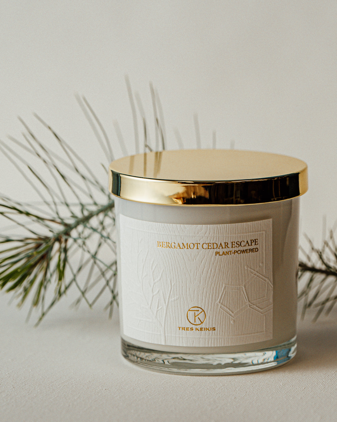 A candle in a clear glass jar with shiny gold lid and a wood-textured white label with embossed plant and molecule art. The words Bergamot Cedar escape and Tres Keikis logo are printed in gold foil. The jar is on a continuous white backdrop with a decorative green pine needle branch behind it.