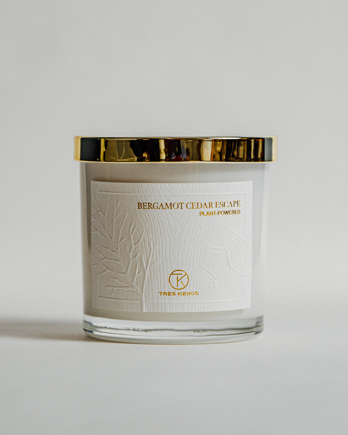 A candle in a clear glass jar with shiny gold lid and a wood-textured white label with embossed plant and molecule art. The words Bergamot Cedar escape and Tres Keikis logo are printed in gold foil. The jar is on a continuous white backdrop.
