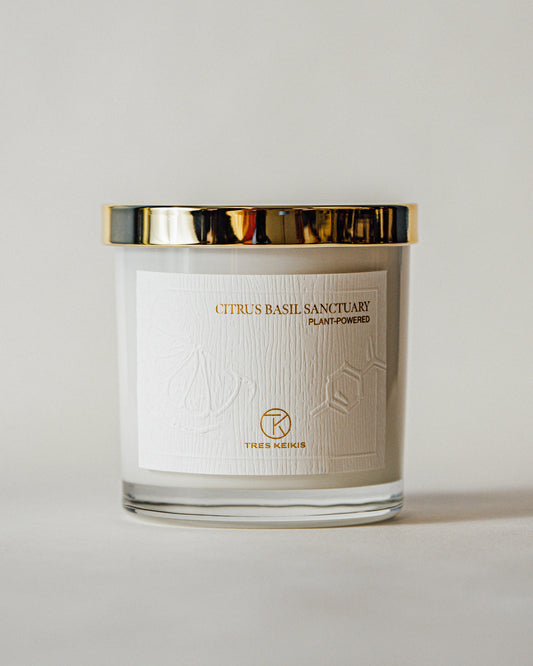 A candle in a clear glass jar with shiny gold lid and a wood-textured white label with embossed plant and molecule art. The words Citrus Basil Sanctuary and Tres Keikis logo are printed in gold foil. The jar is on a continuous white backdrop.