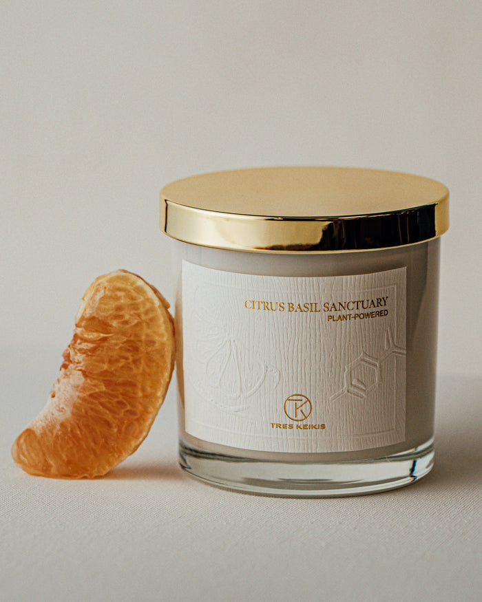 A candle in a clear glass jar with shiny gold lid and a wood-textured white label with embossed plant and molecule art. The words Citrus Basil Sanctuary and Tres Keikis logo are printed in gold foil. The jar is on a continuous white backdrop with a decorative orange slice leaning against it.