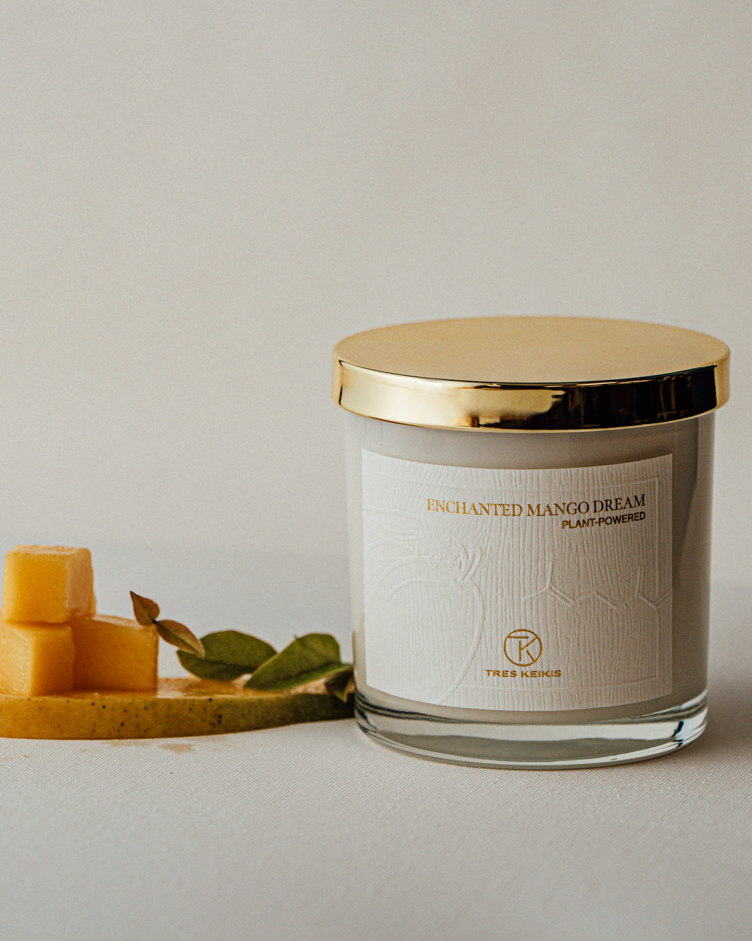 A candle in a clear glass jar with shiny gold lid and a wood-textured white label with embossed plant and molecule art. The words Enchanted Mango Dream and Tres Keikis logo are printed in gold foil. Decorative sliced and cubed mango with green garnish sit beside it and it all is on a white backdrop.