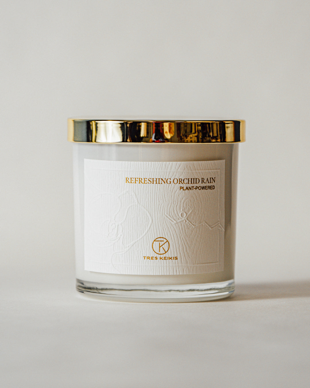 A candle in a clear glass jar with shiny gold lid and a wood-textured white label with embossed plant and molecule art. The words Refreshing Orchid Rain and the Tres Keikis logo are printed in gold foil. The jar is on a continuous white backdrop.