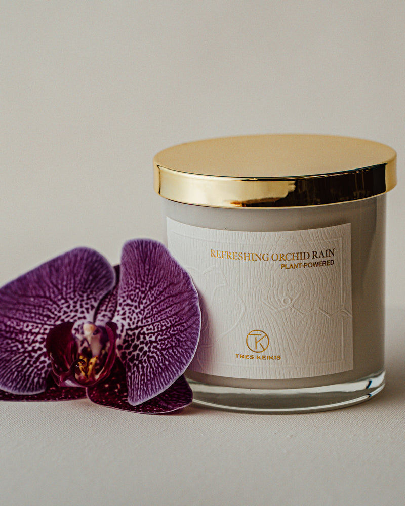 A candle in a clear glass jar with shiny gold lid and a wood-textured white label with embossed plant and molecule art. The words Refreshing Orchid Rain and Tres Keikis logo are printed in gold foil. A. decorative purple orchid flower sits beside it and it all is on a white backdrop.