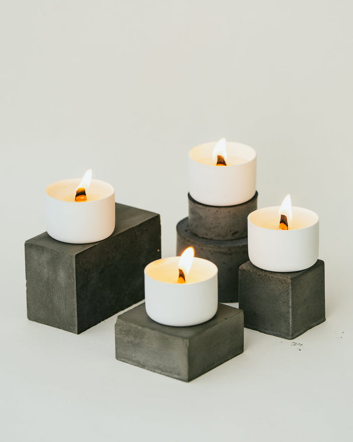 Four lit tealight candles sitting atop small black concrete cubes all atop a white backdrop.